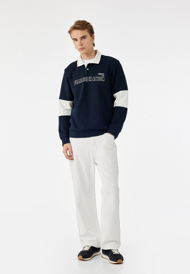 Sudadera azul marino con detalles en crema y texto "SIN MIEDO EN ACCIÓN", combinada con pantalones blancos y zapatillas negras. Diseño de cuello cruzado.