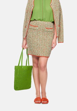 Frau trägt floralen Tweedrock und Jacke über grüner Bluse, hält eine grüne gewebte Tragetasche und trägt orangefarbene gewebte offene Sandalen.