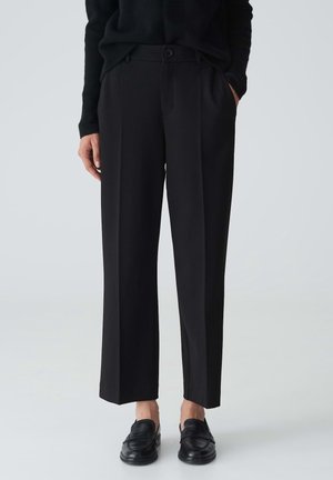 Persoon draagt zwarte rechte broek met knoopsluiting en zwarte instapschoenen, staand tegen een effen lichte achtergrond.