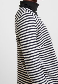 Raidallinen neulepusero musta-valkoisena, jossa on korkea kaulus, vaakasuorat raidat ja teksturoitu kangas. Muotoiltu tiiviiksi istuvuudeksi.