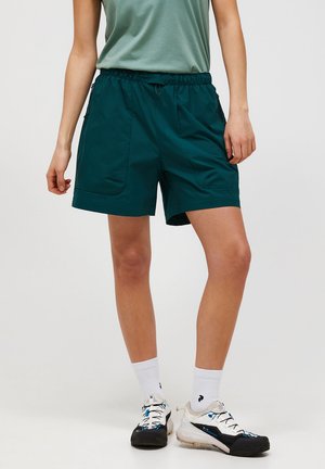 Person iført mørkegrønne løse shorts, lysegrøn ærmeløs top, hvide sokker og hvide-sorte vandresko, stående mod en ensfarvet baggrund.