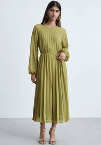Rochie plisată verde măsliniu cu mâneci lungi, material texturat și talie ajustată. Purtați cu sandale aurii. Decolteu rotund simplu.