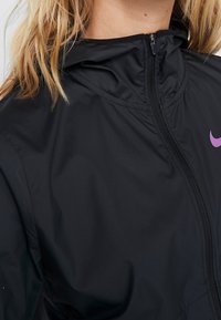 Primer plano de una persona con una chaqueta negra con cremallera y un logo de Nike púrpura cerca del cuello, con el cabello rubio visible.