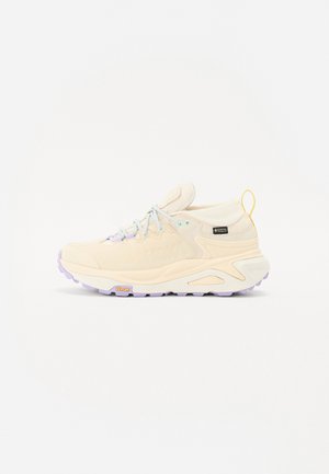 Chaussure de randonnée beige à coupe basse avec semelle épaisse, semelle extérieure Vibram, lacets violet clair, étiquette Gore-Tex et languette de talon jaune.
