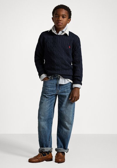 Námornícky pletený sveter s kabelovým vzorom, vrstvený cez svetlomodrú prúžkovanú košelu, spárovaný so svetlomodrými džínsami a hnedými koženými loafermi. Jednoduchý, klasický dizajn.