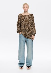 Kardigan w panterkę w kolorze brązowym i czarnym, oversize, z szerokim dekoltem i teksturowanym materiałem. W zestawie z jasnoniebieskimi spodniami typu wide-leg.