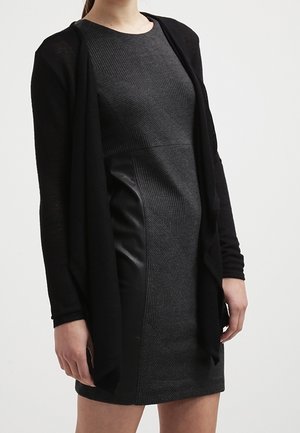 Zwarte cardigan over een aansluitende grijze jurk, met een textuur gebreid bovenstuk en een glad leren paneel aan de zijkant.