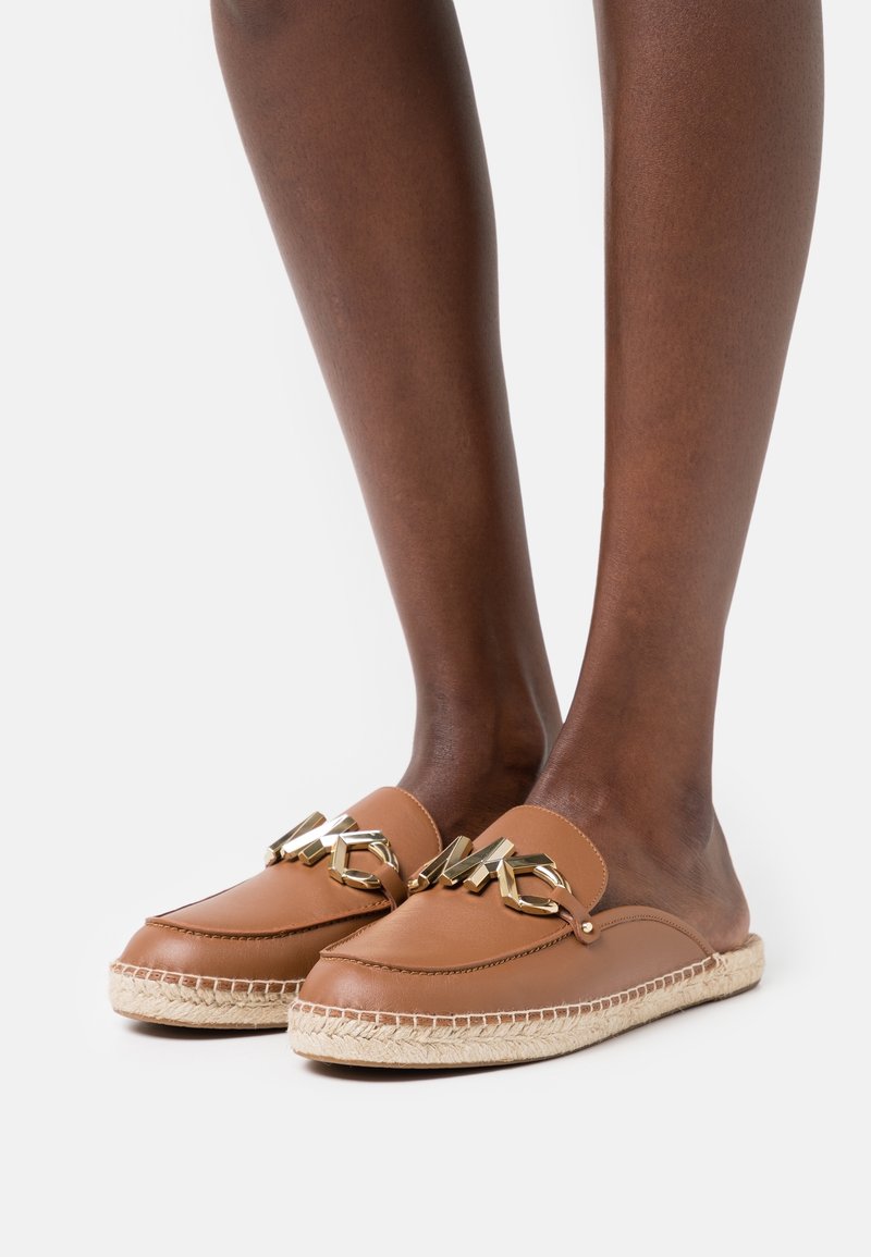 Mocassins marron en cuir à enfiler avec détail chaîne dorée et semelle espadrille tissée portés sur des pieds à la peau foncée sur fond blanc.
