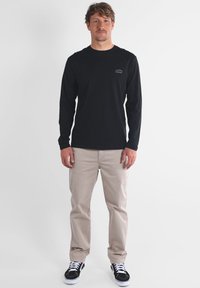 Chemise noire à manches longues avec un petit logo sur la poitrine, associée à un pantalon beige et des baskets noires. Fond uni, posture droite.