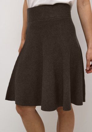 Femme portant une jupe tricotée marron foncé arrivant au genou, avec une taille haute et un léger évasement, accompagnée d'un haut blanc.