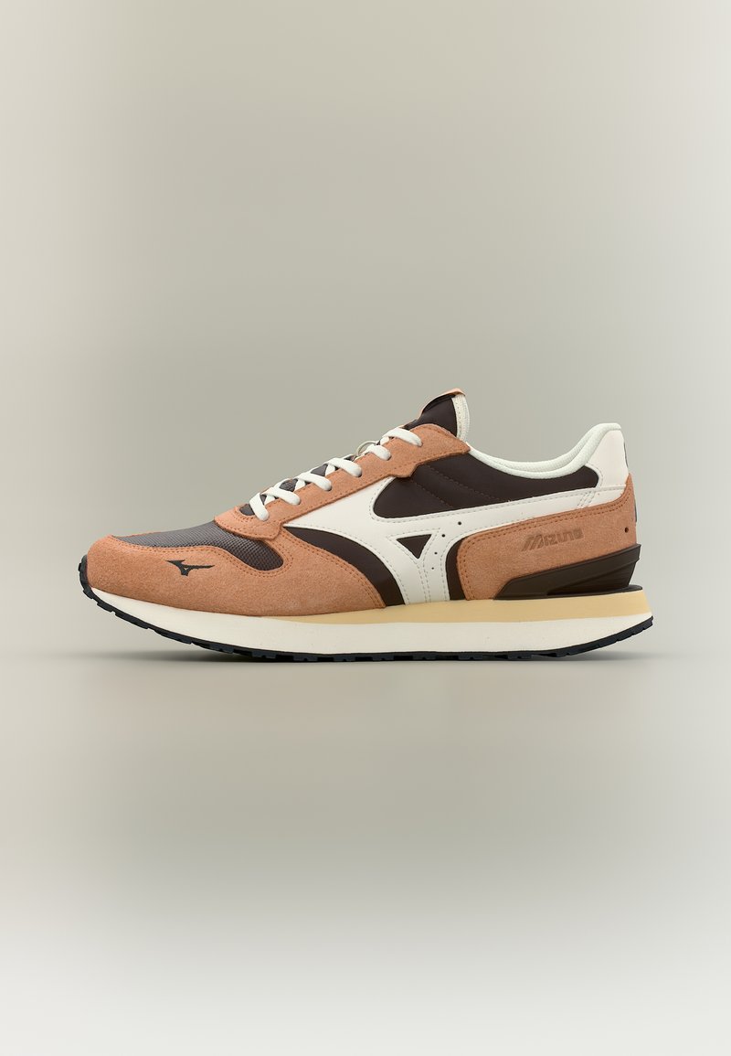 Καφέ, μαύρο και λευκό παπούτσι Mizuno με πάνελ από σουέτ και διχτυωτό υλικό, λευκά κορδόνια και μπεζ σόλα σε απλό μπεζ φόντο.