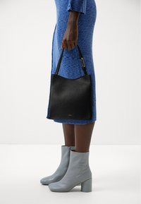 Osoba w niebieskiej, prążkowanej sukience trzyma czarną skórzaną torebkę, nosi jasnoszare botki za kostkę na blokowym obcasie na białym tle.