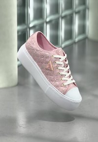 Baskets basses texturées roses avec lacets blancs, semelle blanche épaisse et un logo triangulaire doré sur un fond gris réfléchissant.