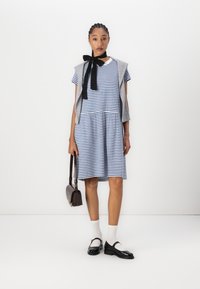 Gestreiftes blau-weißes Kleid mit kurzen Ärmeln, gerafftem Taillenbereich, kombiniert mit einem grauen Sweater-Westen, schwarzer Fliege und schwarzen Schuhen.