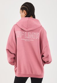 Person med flettehår iført en overdimensjonert rosa hettegenser med teksten "OFF LIMITS DANGEROUS AREA BERLIN 2001" på ryggen, står mot en hvit bakgrunn.