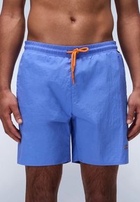 Svjetlomodre kopalke s elastičnim pasom, oranžnim vrvico za zategovanje in majhnim vezenim logotipom na strani. Mehka, teksturirana tkanina.