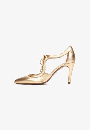 Zapato de cuero dorado con tacón alto, correa en T, punta puntiaguda y laterales recortados. Incluye un lazo decorativo y un tacón delgado y afilado.