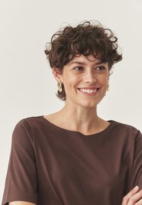 Une personne souriante avec des cheveux courts et bouclés, portant des boucles d'oreilles en or et un haut marron, regardant légèrement sur le côté devant un fond uni.