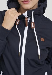 Granatowa kurtka na zamek z kapturem, ozdobiona białymi sznurkami i brązowymi akcentami. Gładka tekstura z elastycznymi mankietami i naszywką z logo.