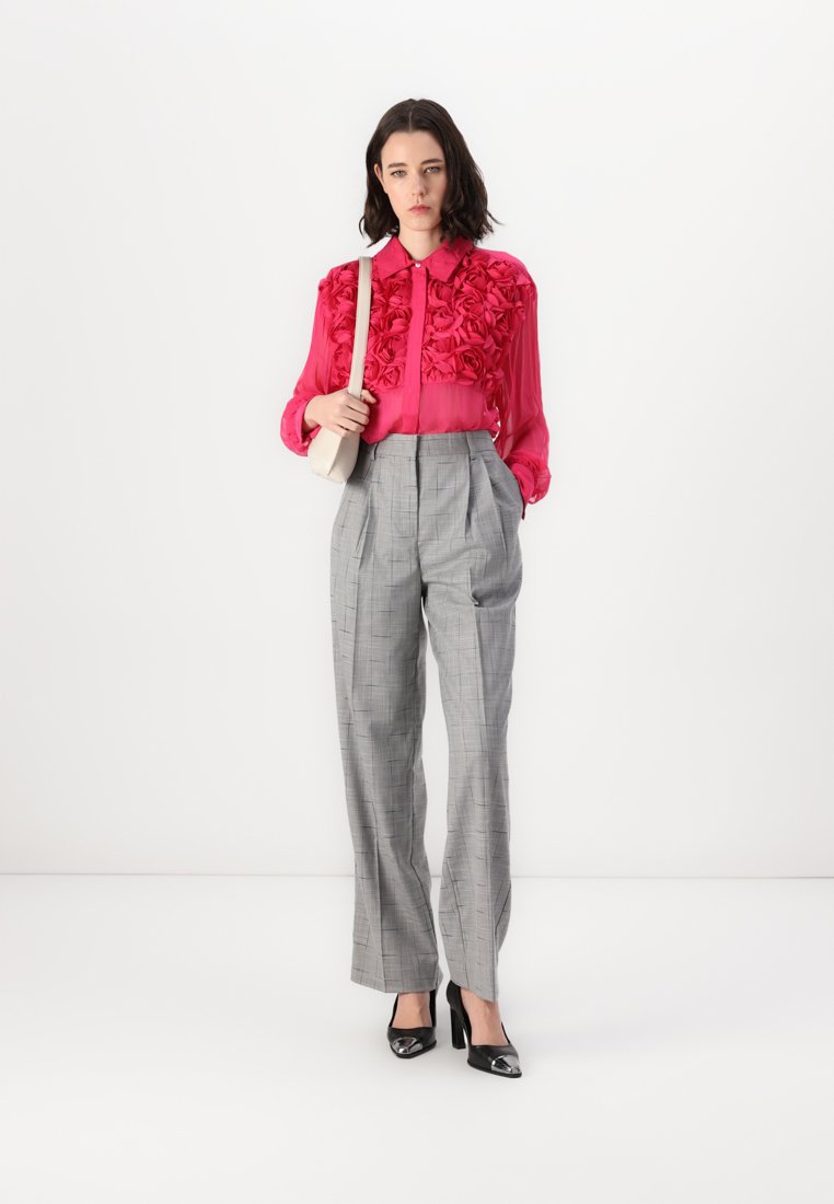 Blouse rose avec des détails floraux, associée à un pantalon taille haute en plaid gris et des escarpins noirs à bout pointu. Sac à main simple porté en bandoulière.
