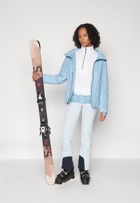 Helles, blaues Skijacke, weißes Reißverschluss-Oberteil, hellblaue Skihose mit marineblauen Akzenten, schwarze Skischuhe, hält braune und schwarze Skier mit Bindungen.