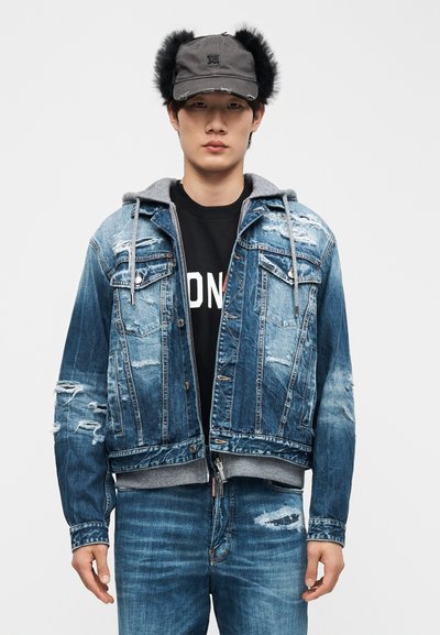 Νεαρός άντρας που φοράει φθαρμένο τζιν μπουφάν με γκρι φούτερ με κουκούλα, μαύρο μπλουζάκι με σχέδιο, σκισμένα τζιν και γκρι καπέλο με μαύρα γούνινα αυτιά.