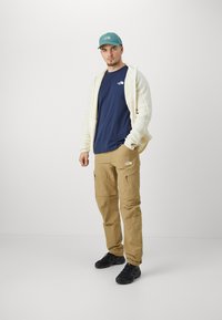 Beige cargo nadrág több cipzáras zsebbel, sötétkék póló, krémszínű kapucnis pulóver és türkiz sapka, fekete túracipővel párosítva.