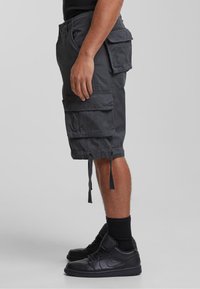 Personne portant un short cargo noir avec de grandes poches et des baskets montantes noires, se tenant devant un fond uni.