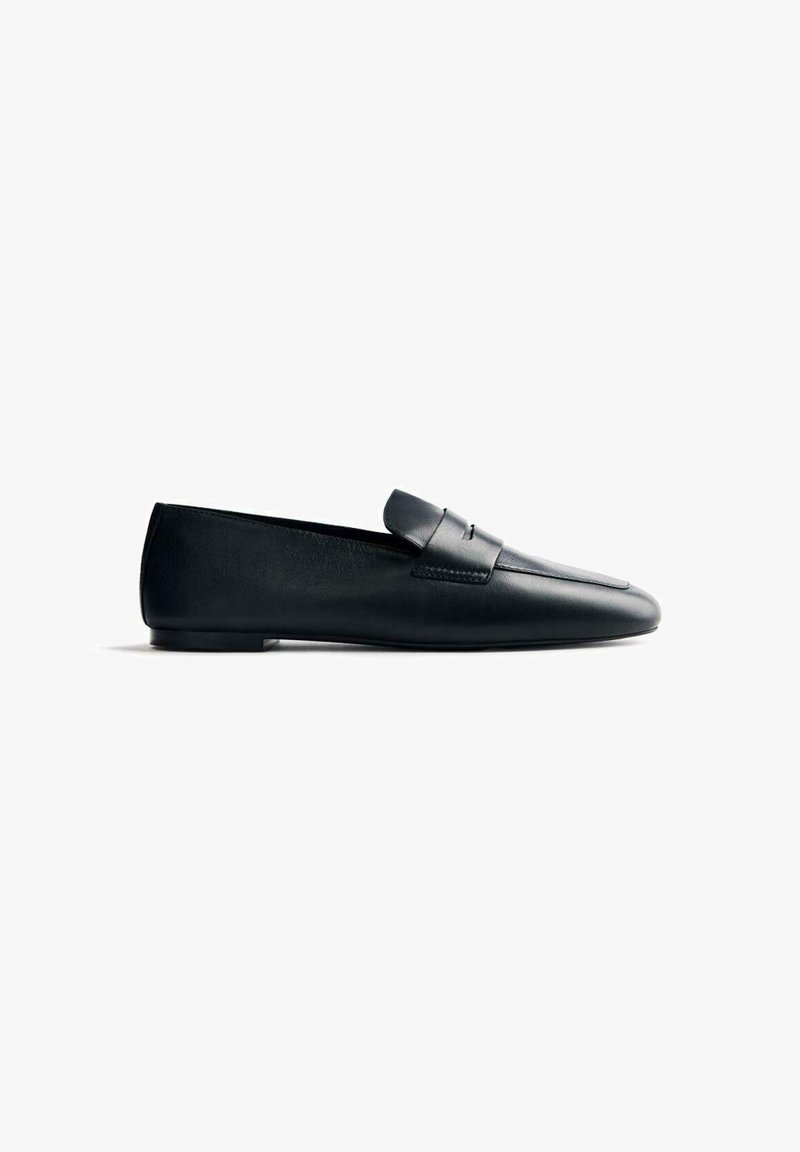 Mocasín de cuero negro con la punta afilada y un detalle de correa en la parte delantera. La suela es plana y lisa, ofreciendo un diseño minimalista sin patrones visibles.