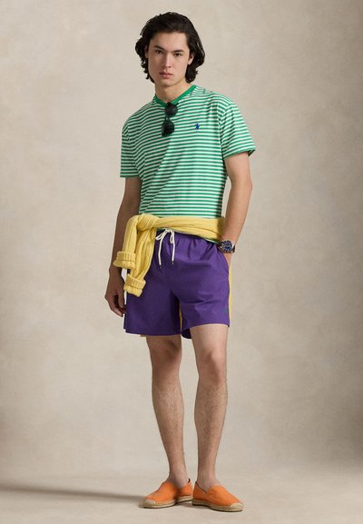 Πράσινο και λευκό ριγέ μπλουζάκι, μοβ σορτς με πλευρική λεπτομέρεια, κίτρινο πουλόβερ δεμένο στη μέση και πορτοκαλί εσπαντρίγιες.