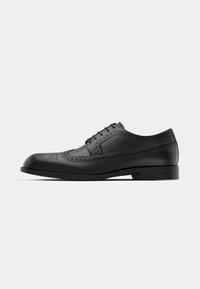 Chaussure de ville brogue pour homme en cuir noir avec perforations en forme d'aile et lacets fermés sur une semelle plate, sur un fond clair uni.