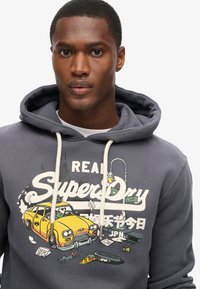Felpa grigia con una stampa grafica che presenta un taxi giallo, elementi cartoon e il testo "REAL SuperDry". Dettagli con coulisse di color crema.