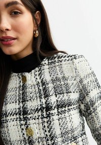 Veste à carreaux noir et blanc avec un design texturé, dotée d'un col montant noir et de détails de boutons dorés.
