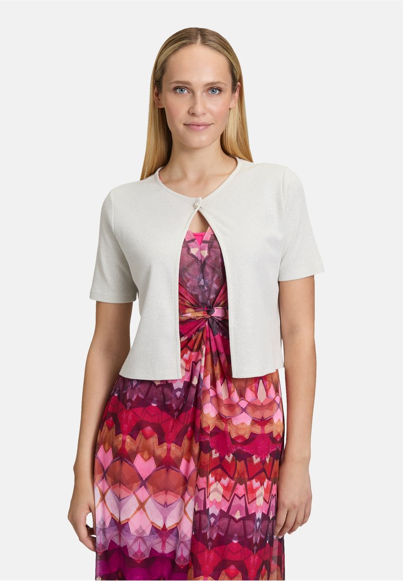 Witte kort mouwen cardigan met een ronde hals en een sluiting aan de voorkant. Draagt over een roze en rode geometrisch patroon jurk.