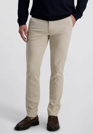 Man draagt beige slim-fit broek met zwarte trui en donkerbruine leren loafers, staand met één hand in de zak.