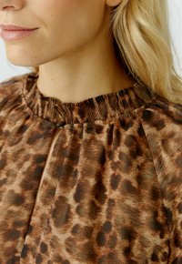 Bruine blouse met luipaardprint, voorzien van een gerimpelde halslijn en een sleutelgatdetail. De stof is lichtgewicht met een gladde textuur.