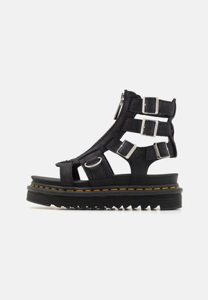 Sandalias de plataforma de cuero negro con múltiples correas ajustables, hebillas metálicas y una suela de goma texturizada con un detalle de costura amarilla.