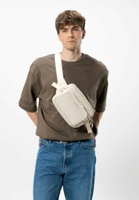 Borsa a tracolla beige realizzata in materiale liscio, con forma rettangolare, chiusura con zip e tracolla regolabile, indossata su una camicia marrone.