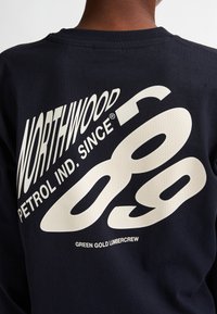 Felpa blu scuro con design di testo bianco: "NORTHWOOD" e "PETROL IND. DAL 89" in stili diversi, con dettagli testurizzati sui numeri.
