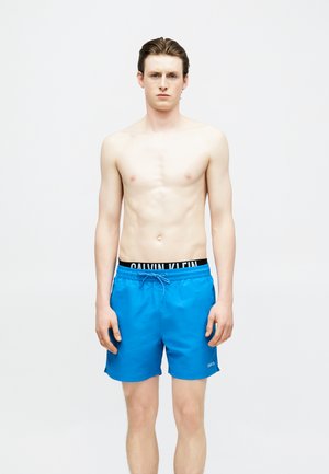 Fiatal férfi, nedves, hátrafésült hajjal, kék fürdőnadrágot visel, látható fekete Calvin Klein derékpánttal, egyszerű világos háttér előtt áll.