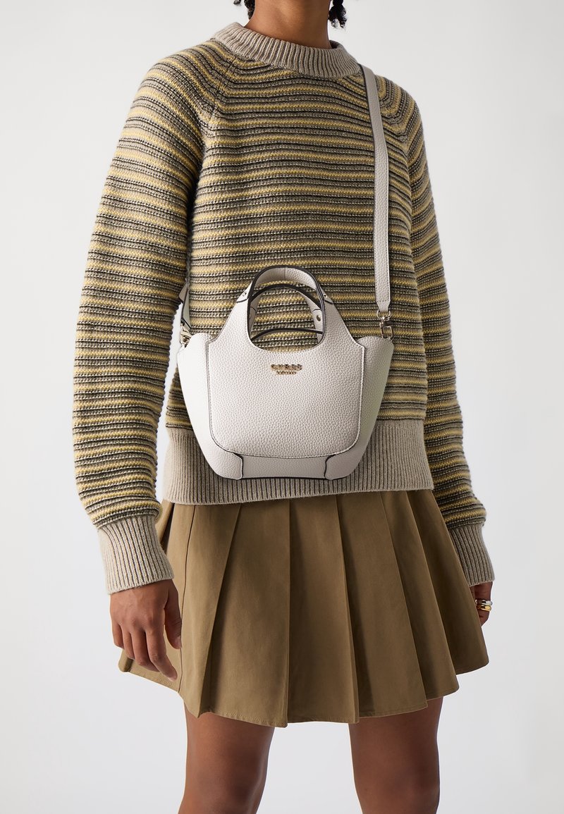 Personne portant un pull rayé et une jupe plissée marron, portant un petit sac à main en cuir blanc texturé avec poignées supérieures et une bandoulière.