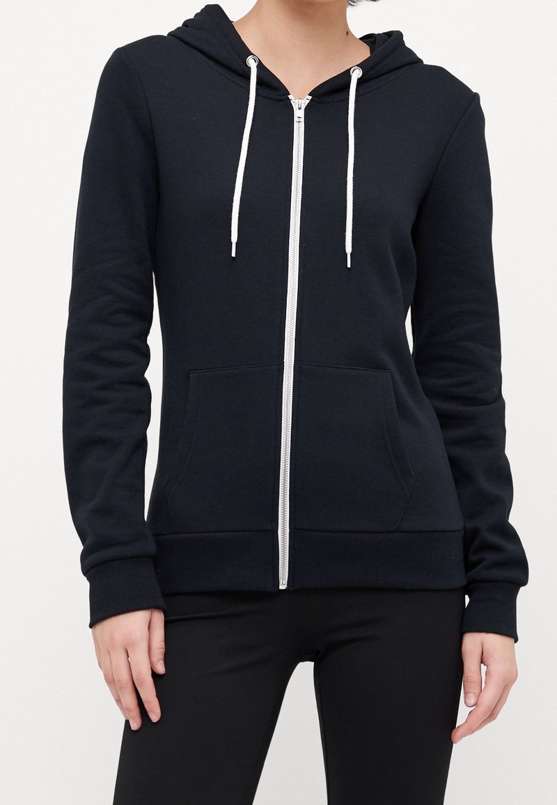 Femme portant un sweat à capuche noir zippé avec des cordons blancs, des poches avant et un pantalon noir, sur un fond blanc uni.