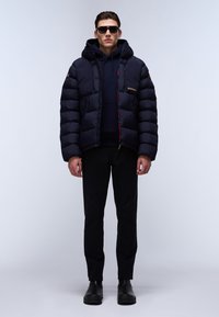 Giacca puffer blu navale con cappuccio, caratterizzata da sezioni trapuntate e dettagli interni rossi, indossata sopra un felpa scura e pantaloni neri. Occhiali da sole neri.