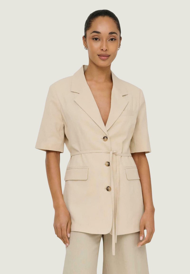 Femme portant un blazer beige à manches courtes ceinturé avec trois boutons et un pantalon assorti, debout devant un fond clair uni.