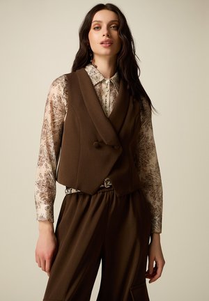 Donna che indossa un gilet marrone su misura sopra una camicia beige a maniche lunghe con motivi floreali e pantaloni marroni a vita alta coordinati, in piedi davanti a uno sfondo semplice.