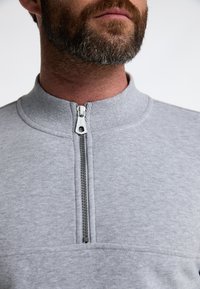 Grå zip-up sweatshirt med høj krave, forsynet med en metallynlås. Stoffet er blødt med en glat overflade og minimale syninger.