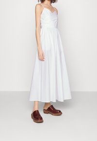 Weißes, ärmelloses Maxikleid mit figurbetontem Oberteil und gerafftem Taillenbund, kombiniert mit bordeauxfarbenen Holzclogs mit rundem Zeh und offener Ferse.
