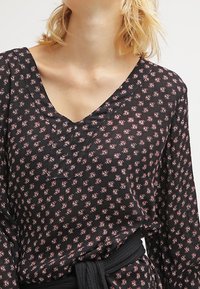 Robe noire avec un petit motif floral rose, décolleté en V, à manches longues et taille cintrée avec une ceinture noire. Tissu doux et léger.