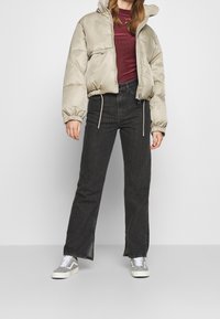 Uppblåsbar jacka i ljusbeige, ribbad mörkröd tröja, mörka raka jeans och grå sneakers med vita detaljer. Dragkedja och snörningsdetaljer.