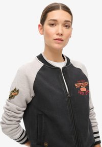 Veste bomber noire et grise avec fermeture éclair à l'avant, poignets côtelés et logo brodé. Comprend un patch en forme d'aile sur la manche gauche.